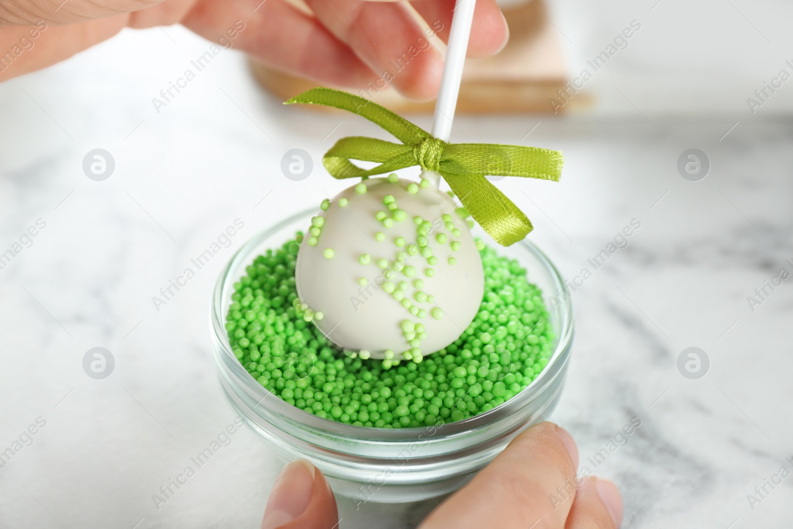 Photo of Young woman putting cake pop into green sprinkles at white marble table, closeup