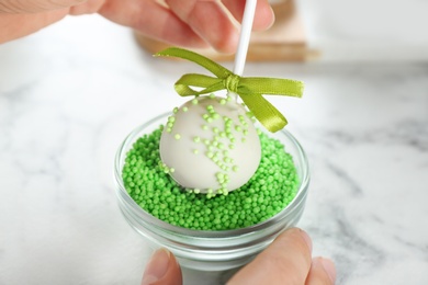 Young woman putting cake pop into green sprinkles at white marble table, closeup