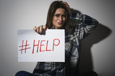 Crying young woman with hashtag HELP near white wall. Domestic violence concept