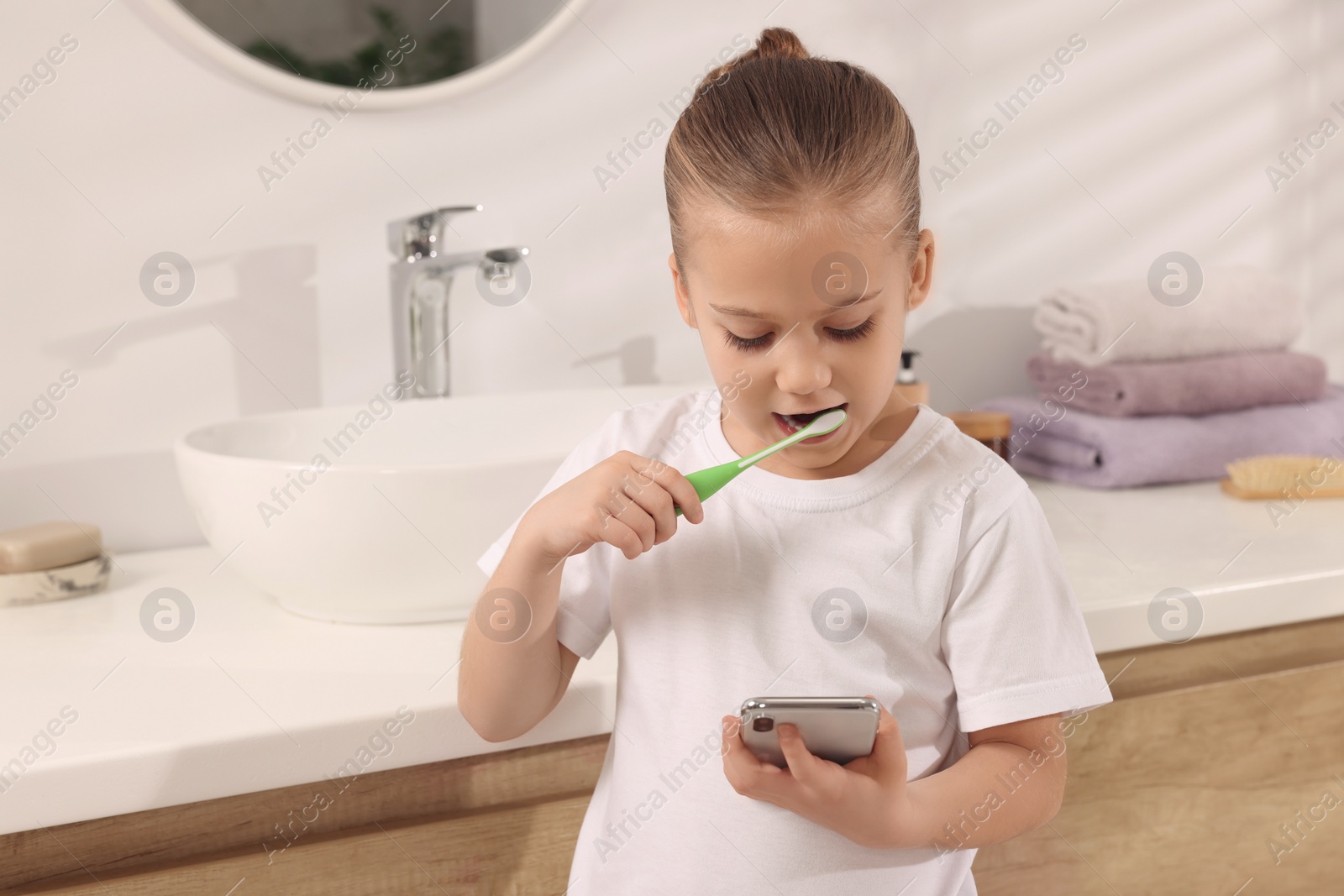 Photo of Little girl using smartphone while brushing teeth in bathroom, space for text. Internet addiction