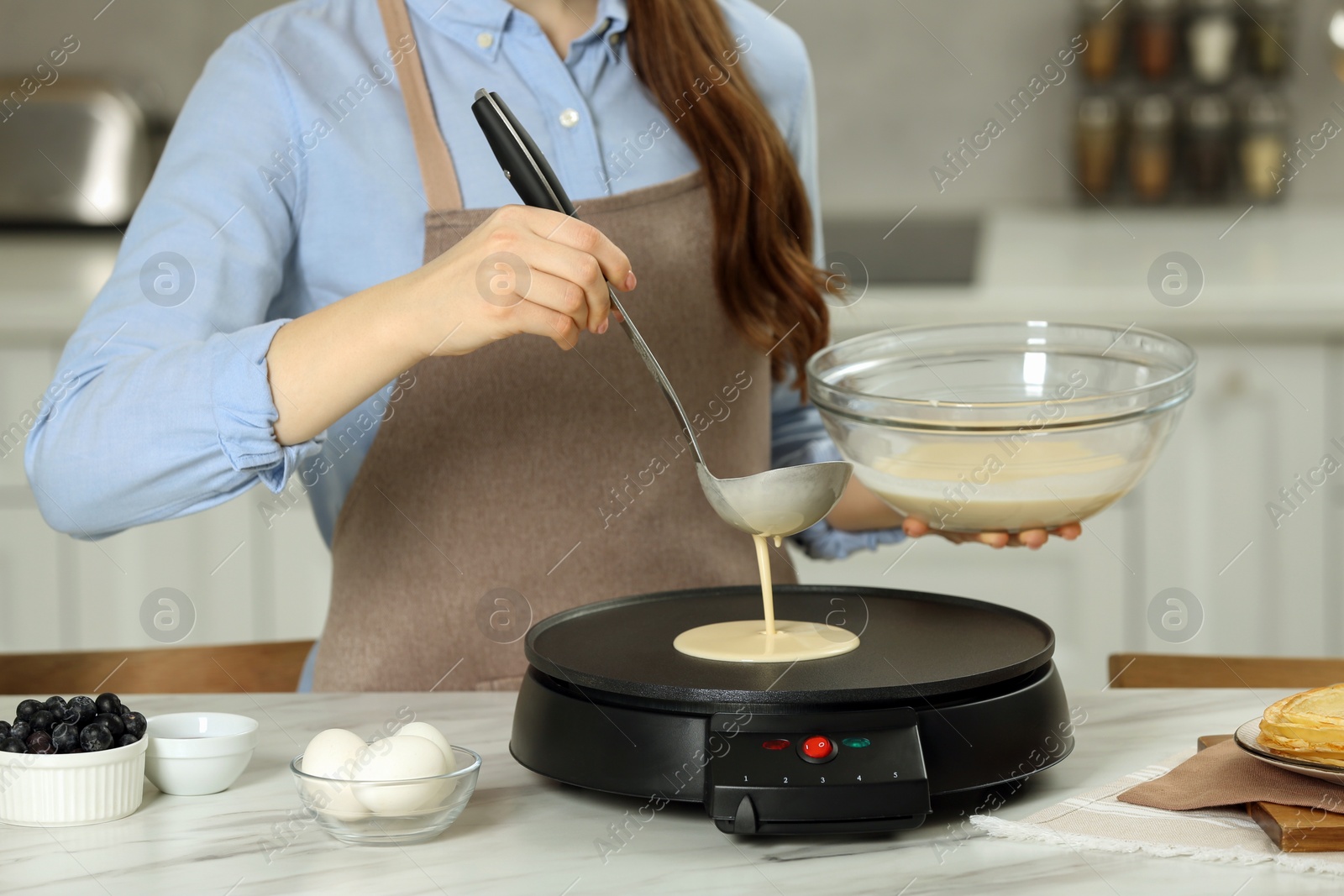 Photo of Woman cooking delicious crepe on electric maker at white marble table in kitchen, closeup