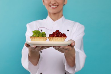 Happy professional confectioner in uniform holding plate with delicious tartlets on light blue background, closeup