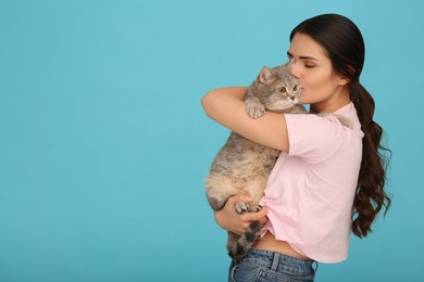 Young woman kissing her adorable cat on light blue background, space for text