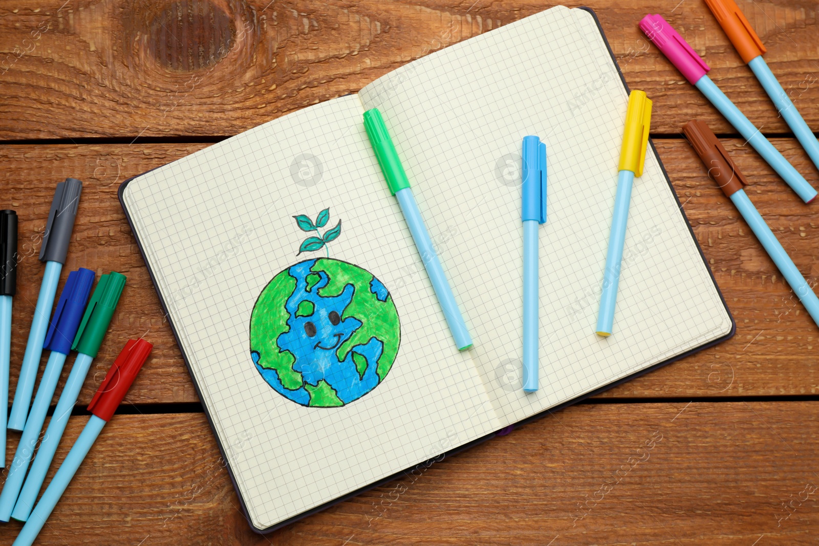 Photo of Happy Earth Day. Notebook with drawing of planet and markers on wooden table, flat lay