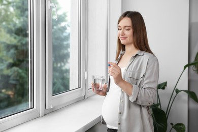 Photo of Beautiful pregnant woman holding pill and glass of water near window at home