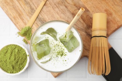 Photo of Glass of tasty iced matcha latte, bamboo whisk and powder on white tiled table, flat lay