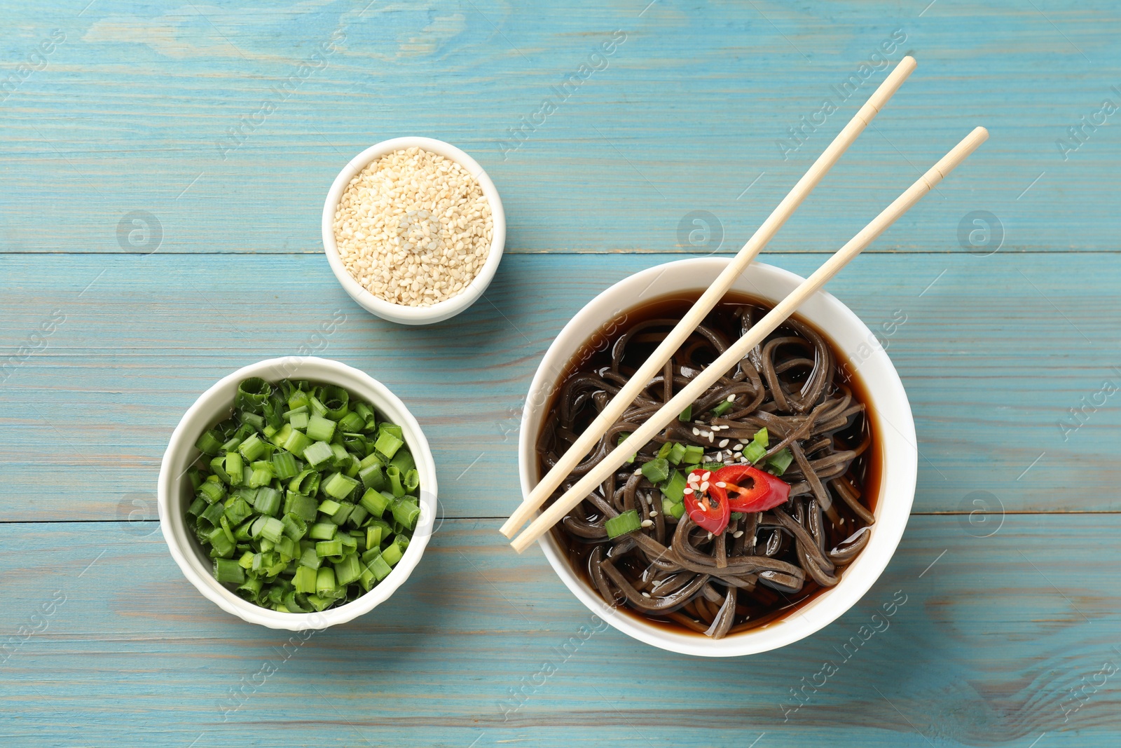 Photo of Tasty buckwheat noodle (soba) soup with chili pepper, green onion, sesame and chopsticks on light blue wooden table, flat lay