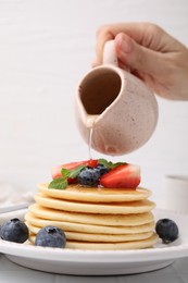 Photo of Woman pouring honey from jug onto delicious pancakes with strawberries, blueberries and mint at white tiled table, closeup