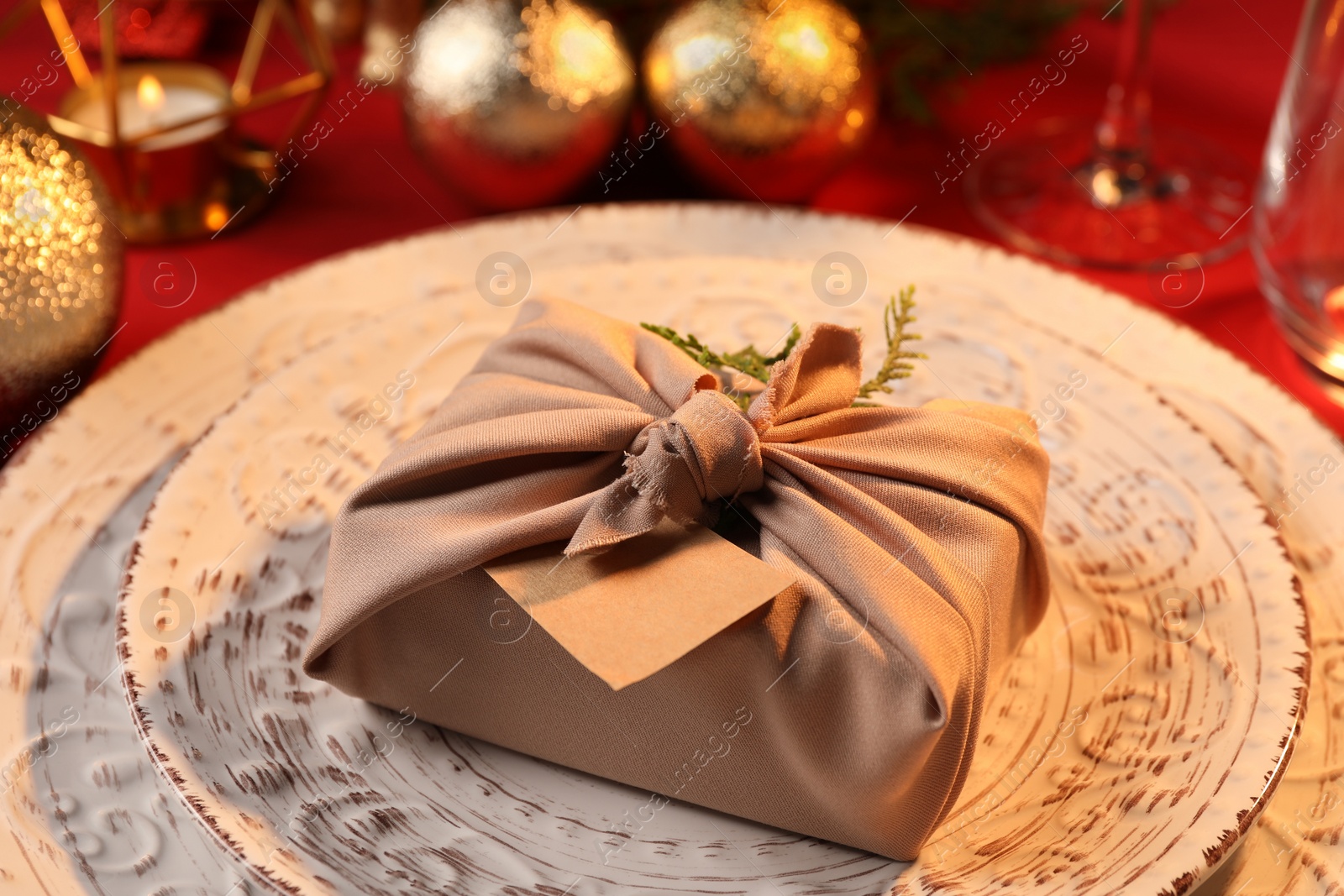 Photo of Furoshiki technique. Gift packed in beige fabric, blank card and festive decor on red table