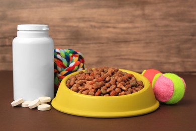Bowl with dry pet food, bottle of vitamins and toys on brown surface