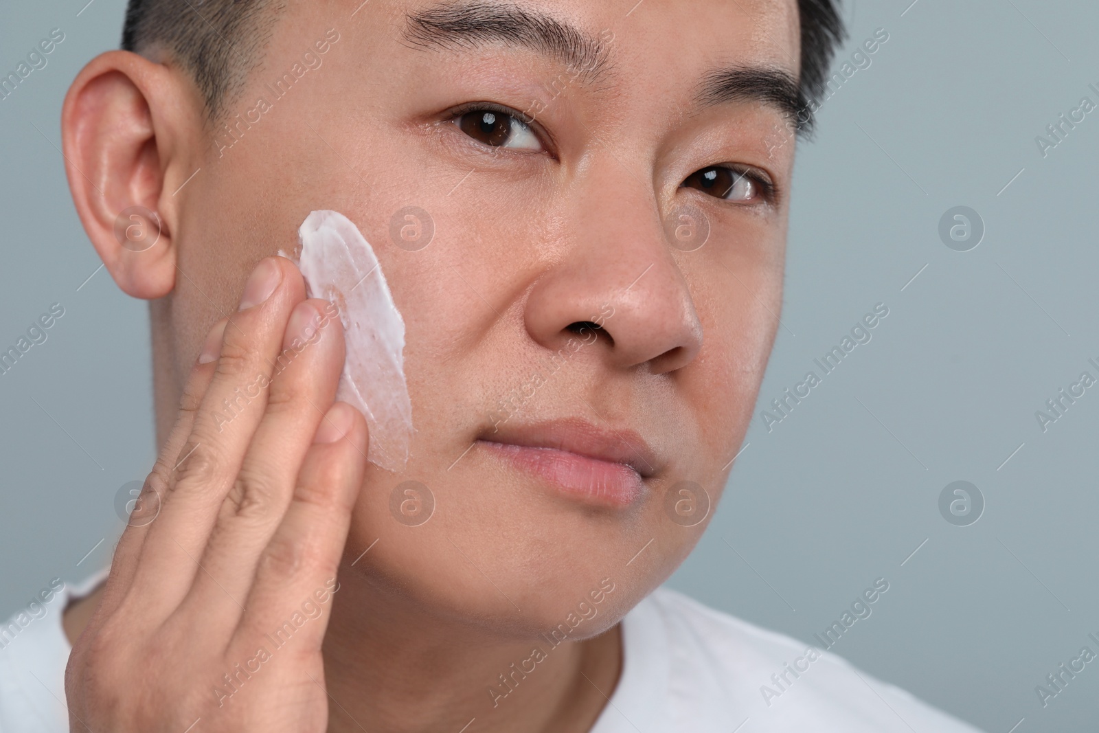Photo of Handsome man applying cream onto his face on light grey background, closeup