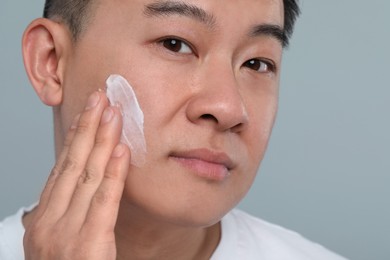 Handsome man applying cream onto his face on light grey background, closeup