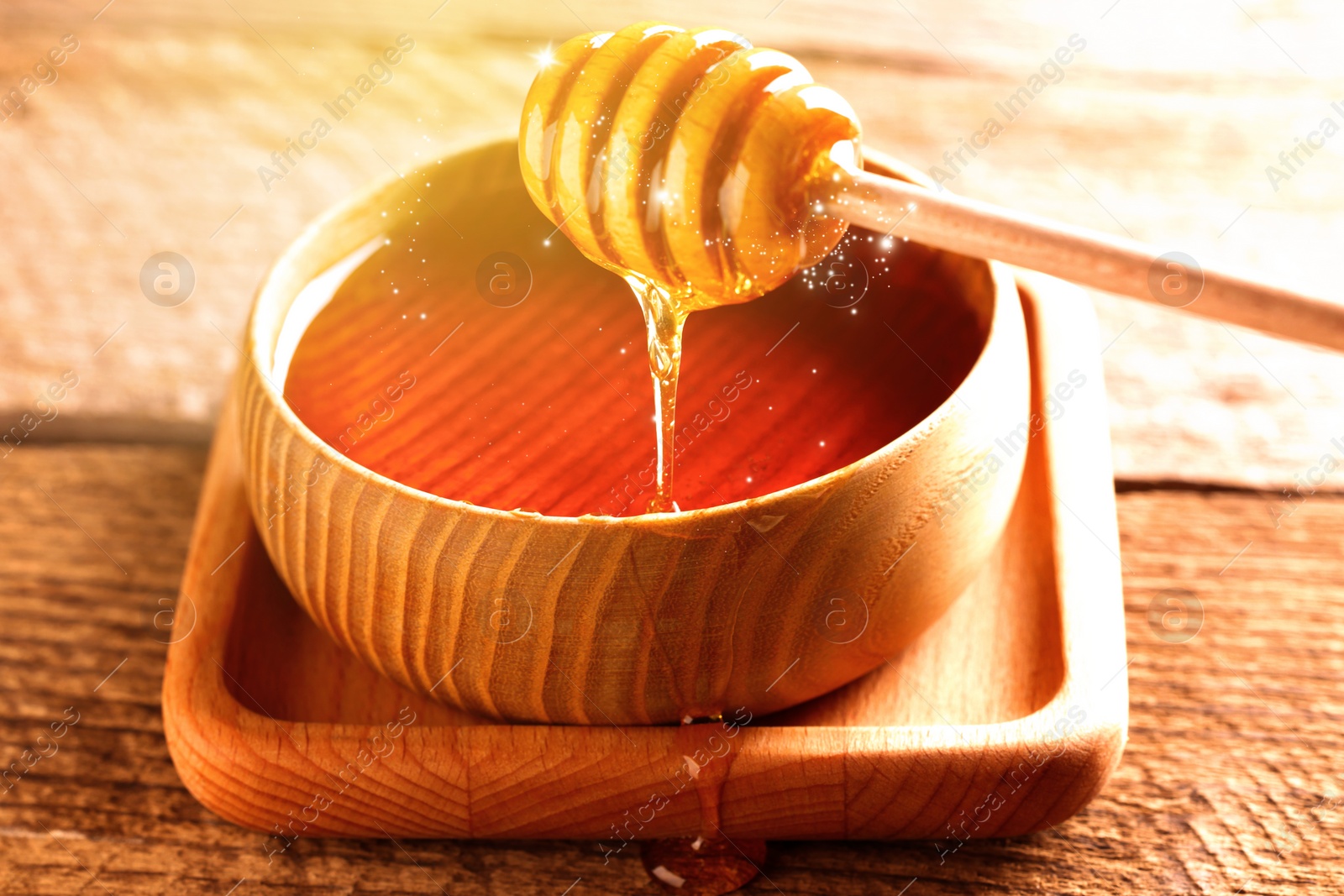 Image of Natural honey dripping from dipper into wooden bowl on table under sunlight, closeup