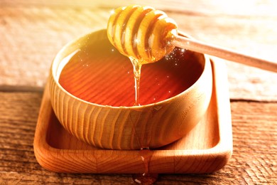 Natural honey dripping from dipper into wooden bowl on table under sunlight, closeup