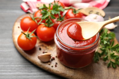 Jar and spoon with tasty ketchup, fresh tomatoes, parsley and spices on grey wooden table, closeup