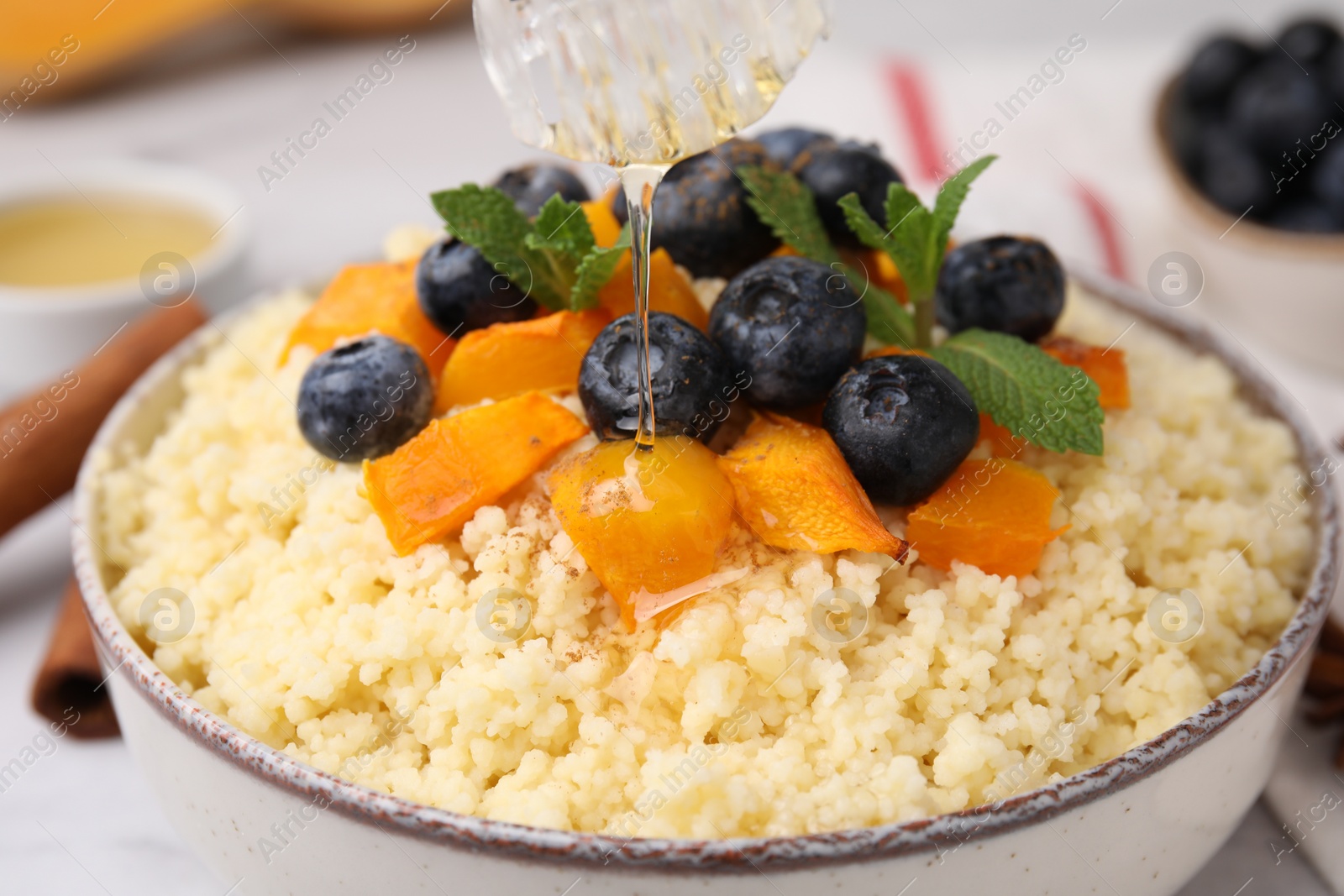 Photo of Pouring honey onto tasty couscous with blueberries, pumpkin and mint, closeup