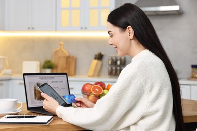 Photo of Beautiful young woman with credit card and gadgets shopping online at wooden table in kitchen