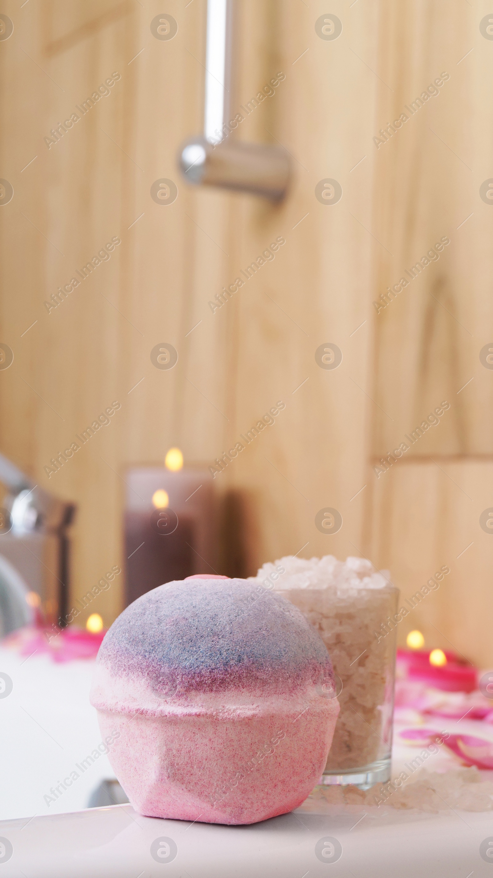 Photo of Colorful bath bomb, sea salt, flower petals and burning candles on white tub in bathroom