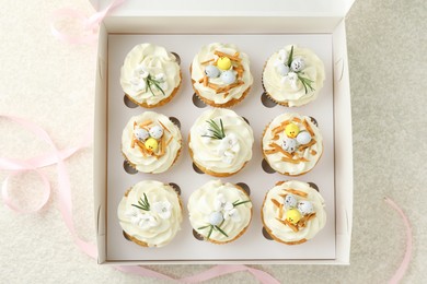 Tasty Easter cupcakes in box and pink ribbon on beige table, flat lay