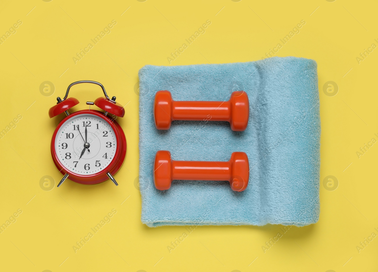 Photo of Alarm clock, towel and dumbbells on yellow background, flat lay. Morning exercise