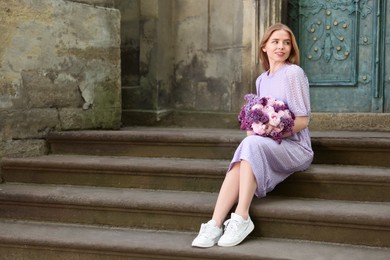 Photo of Beautiful woman with bouquet of spring flowers on stairs near building, space for text