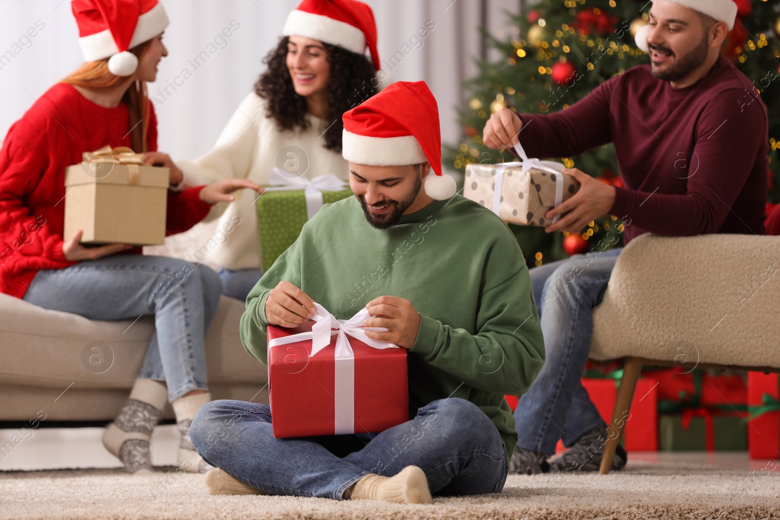 Photo of Christmas celebration in circle of friends. Happy young man opening gift at home, selective focus