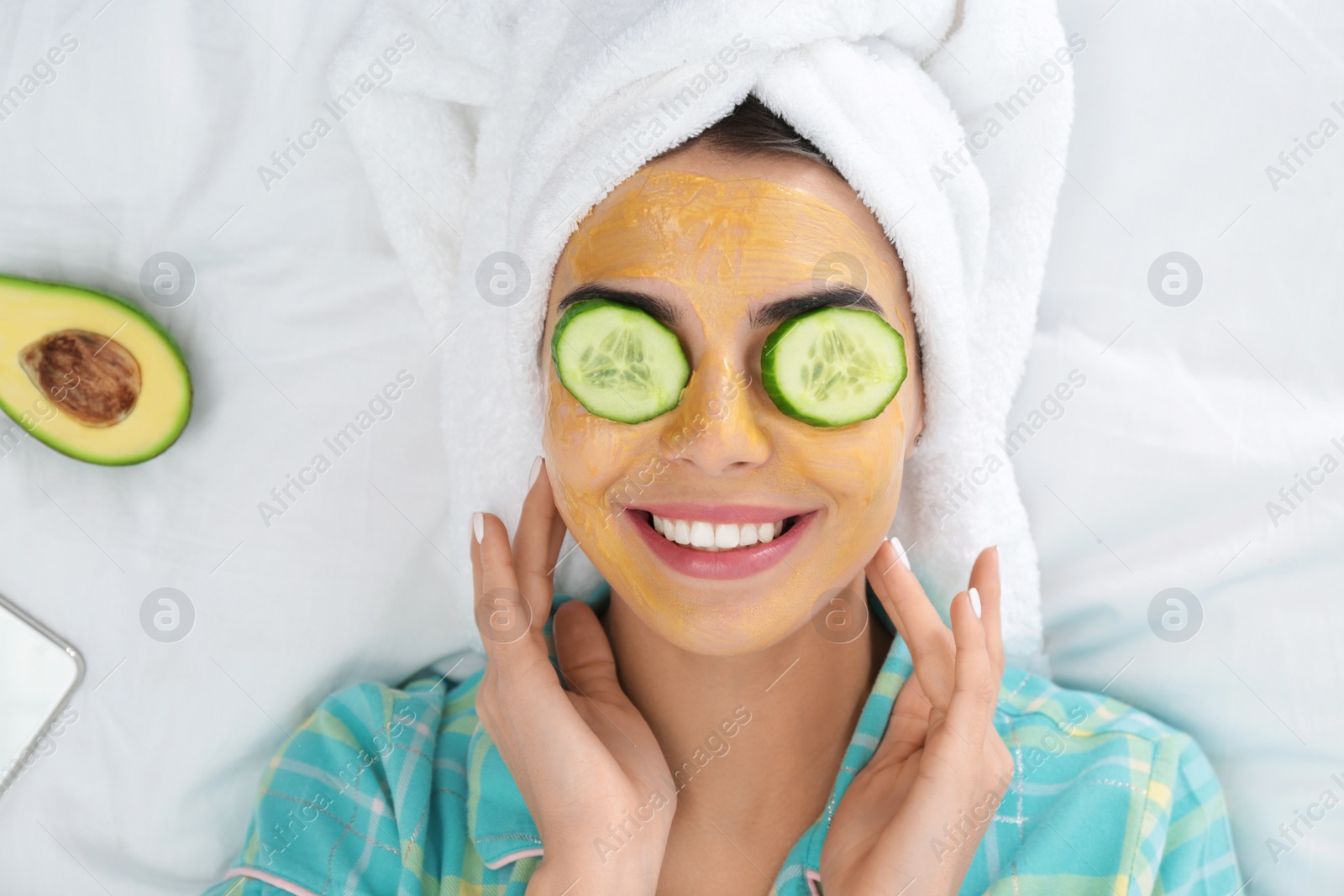 Photo of Young woman with facial mask and cucumber slices lying on bed, top view