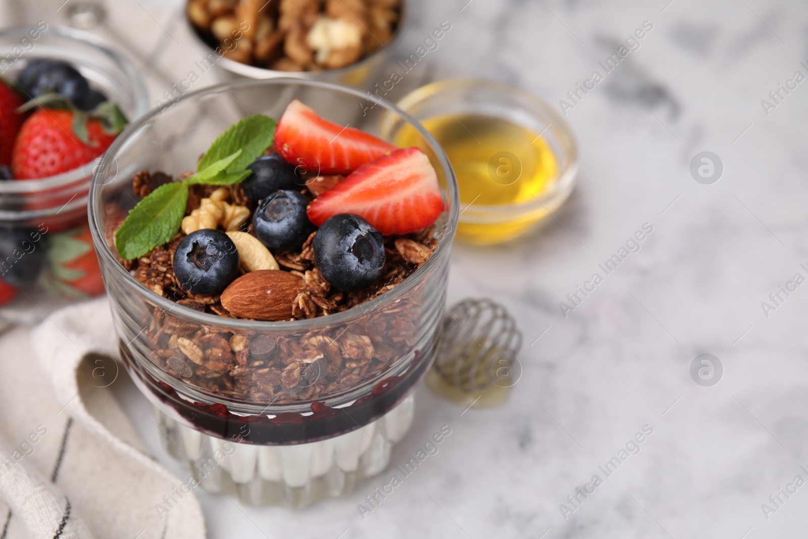 Photo of Tasty granola with berries, nuts, jam and yogurt in glass on white marble table, closeup. Space for text