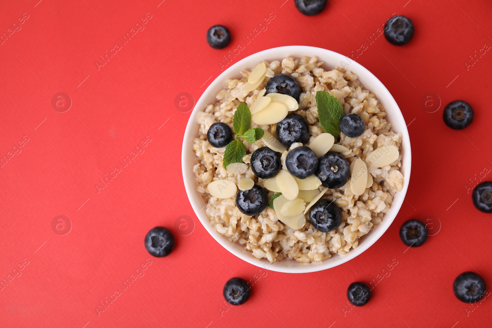 Photo of Tasty oatmeal with blueberries, mint and almond petals in bowl surrounded by fresh berries on red background, flat lay. Space for text