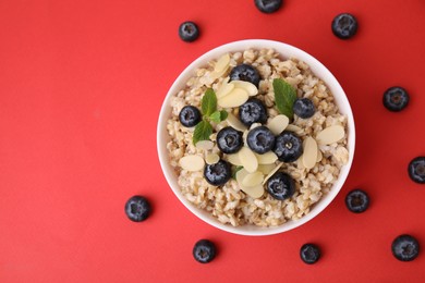 Photo of Tasty oatmeal with blueberries, mint and almond petals in bowl surrounded by fresh berries on red background, flat lay. Space for text