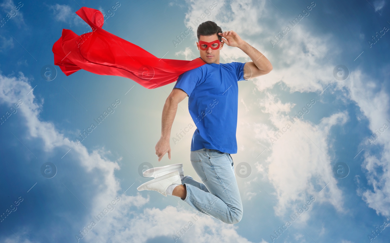 Image of Man in superhero cape and mask jumping against blue sky with clouds