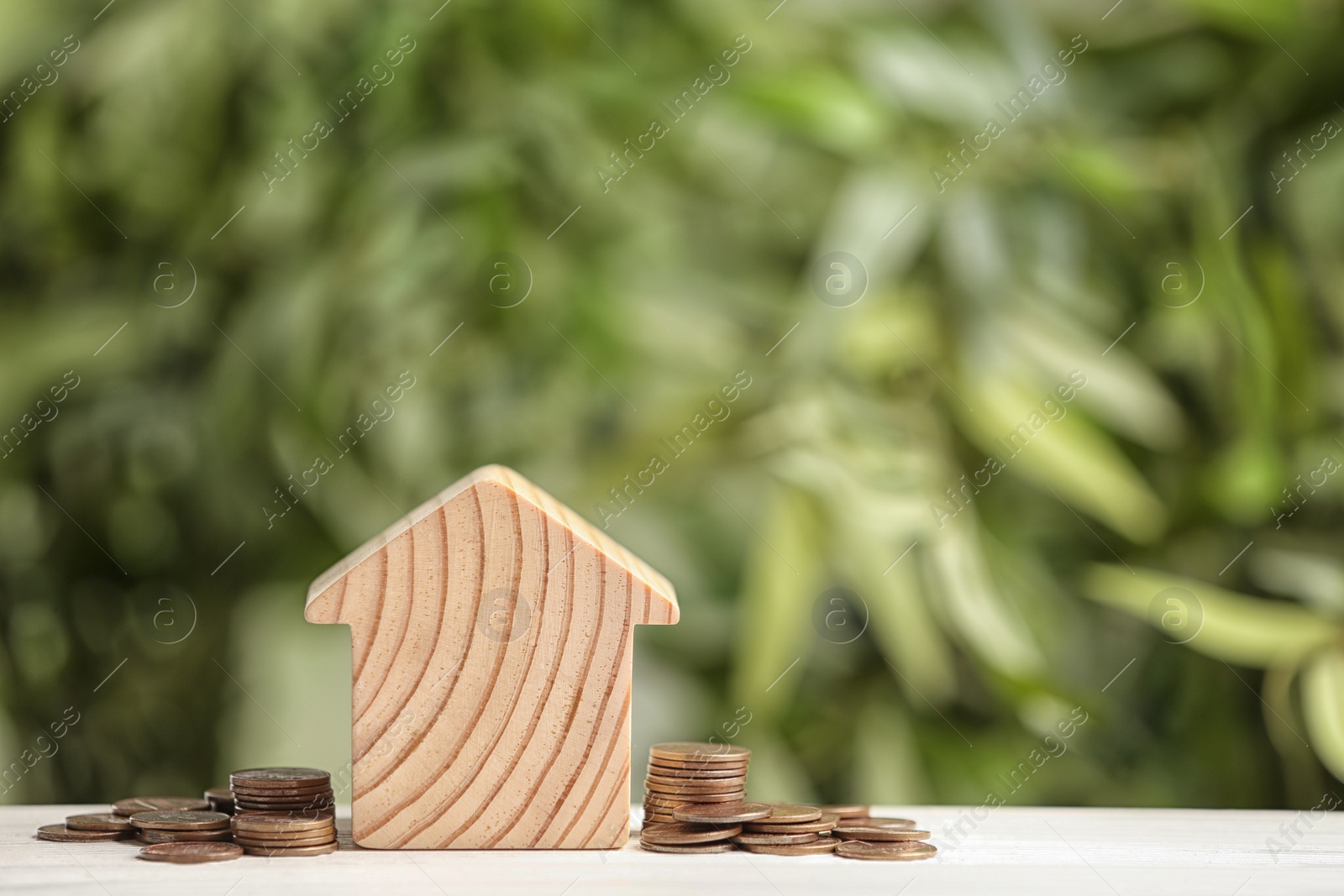 Photo of House model and coins on white table against blurred green background. Space for text