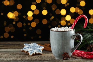 Photo of Delicious hot chocolate with marshmallows and candy cane on wooden table against blurred lights, space for text