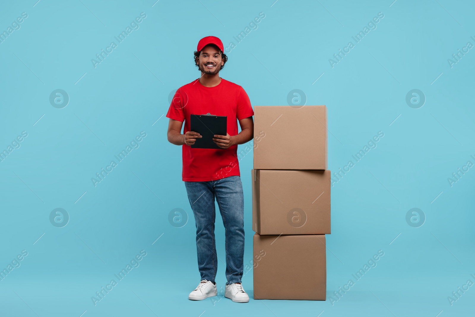 Photo of Happy young courier with clipboard and stack of parcels on light blue background, space for text
