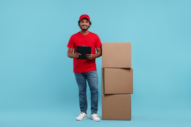 Happy young courier with clipboard and stack of parcels on light blue background, space for text