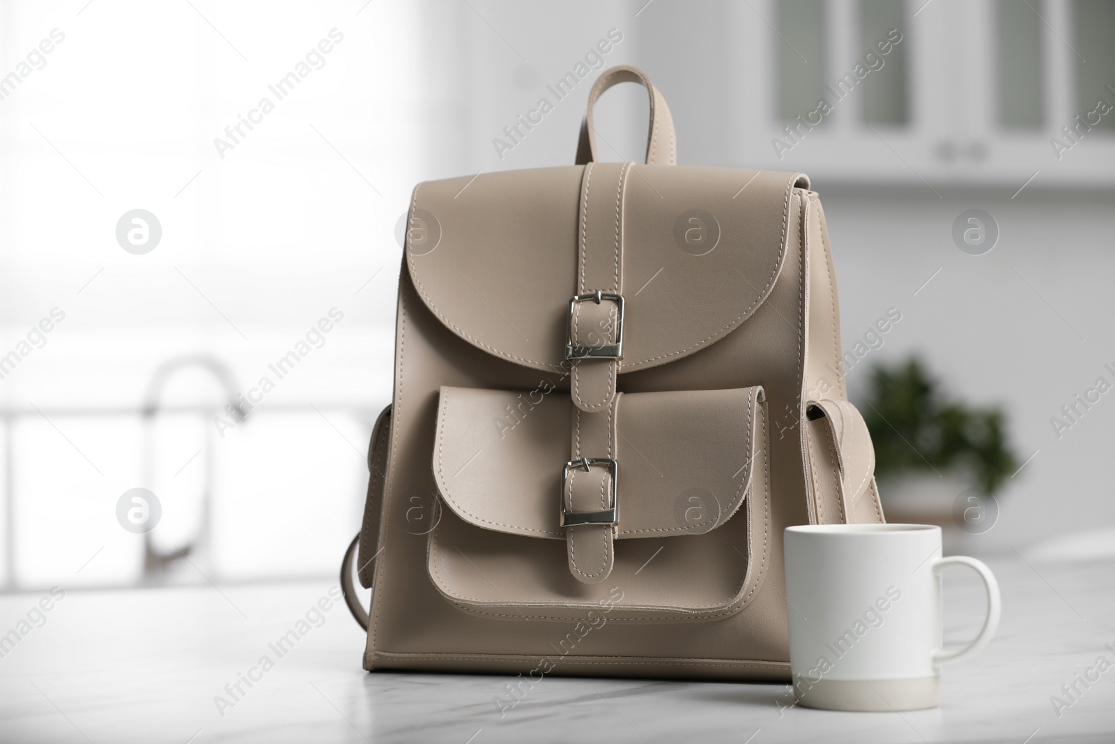 Photo of Stylish beige backpack and cup of coffee on white table in kitchen