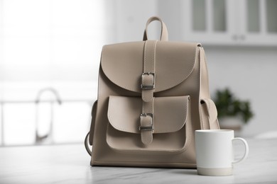 Stylish beige backpack and cup of coffee on white table in kitchen