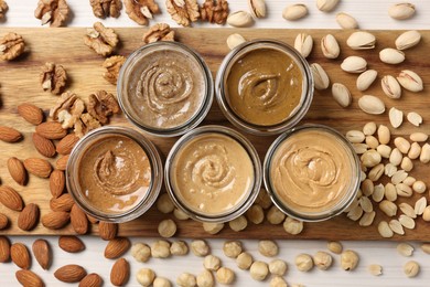 Many tasty nut butters in jars and nuts on white table, flat lay