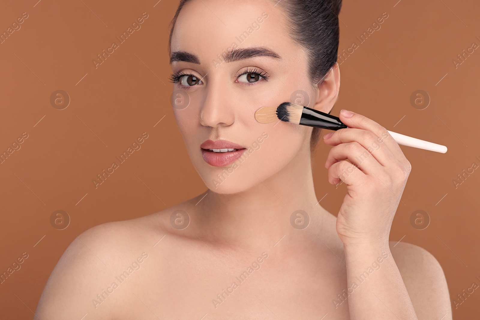 Photo of Woman applying foundation on face with brush against brown background