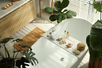 Photo of Bath tray with spa products, towels and shells on tub in bathroom, above view. Interior design