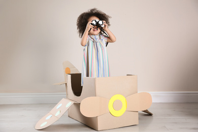 Photo of Cute African American child playing with cardboard plane and binoculars near beige wall