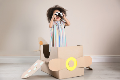 Cute African American child playing with cardboard plane and binoculars near beige wall