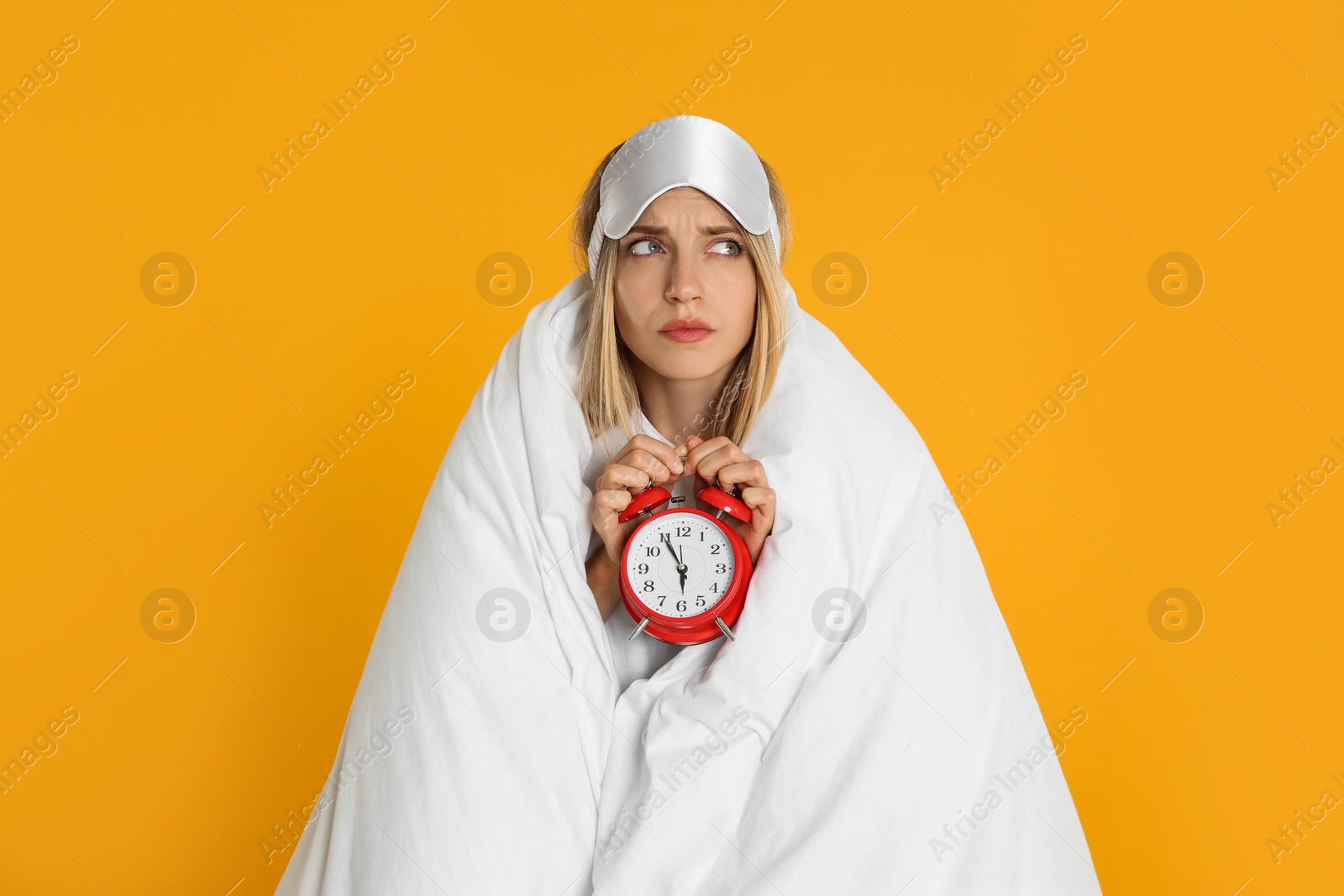 Photo of Sad young woman in sleeping mask wrapped with blanket holding alarm clock on yellow background