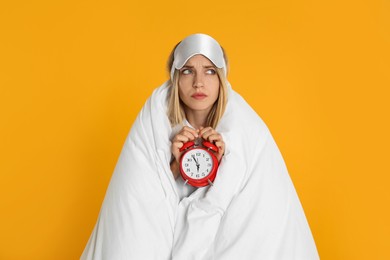 Sad young woman in sleeping mask wrapped with blanket holding alarm clock on yellow background