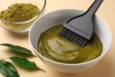 Photo of Bowl of henna cream, brush and green leaves on beige background, closeup. Natural hair coloring