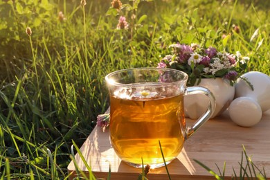 Photo of Cup of aromatic herbal tea, pestle and ceramic mortar with different wildflowers on green grass outdoors