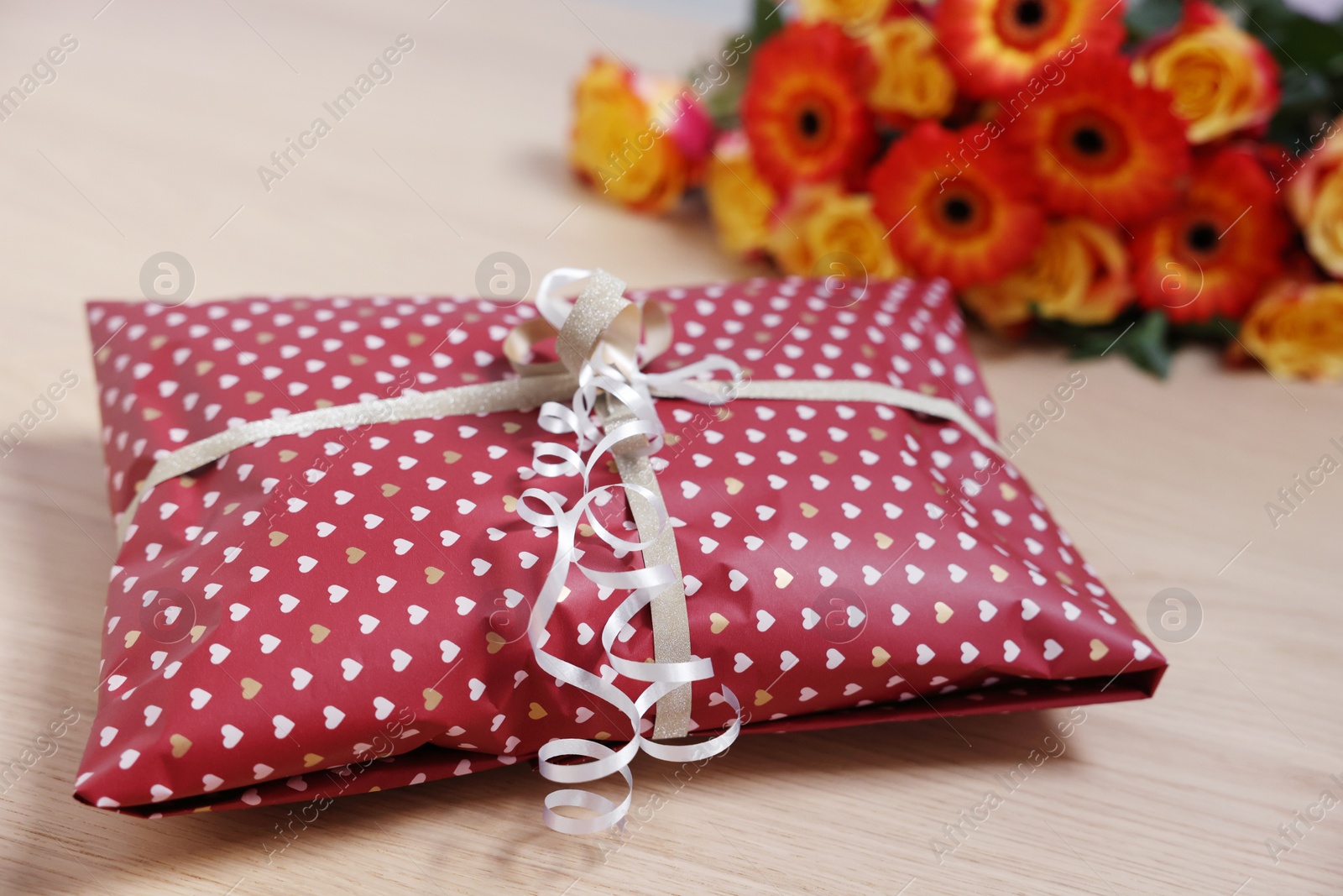 Photo of Parcel wrapped in heart patterned paper and beautiful flowers on wooden table, closeup