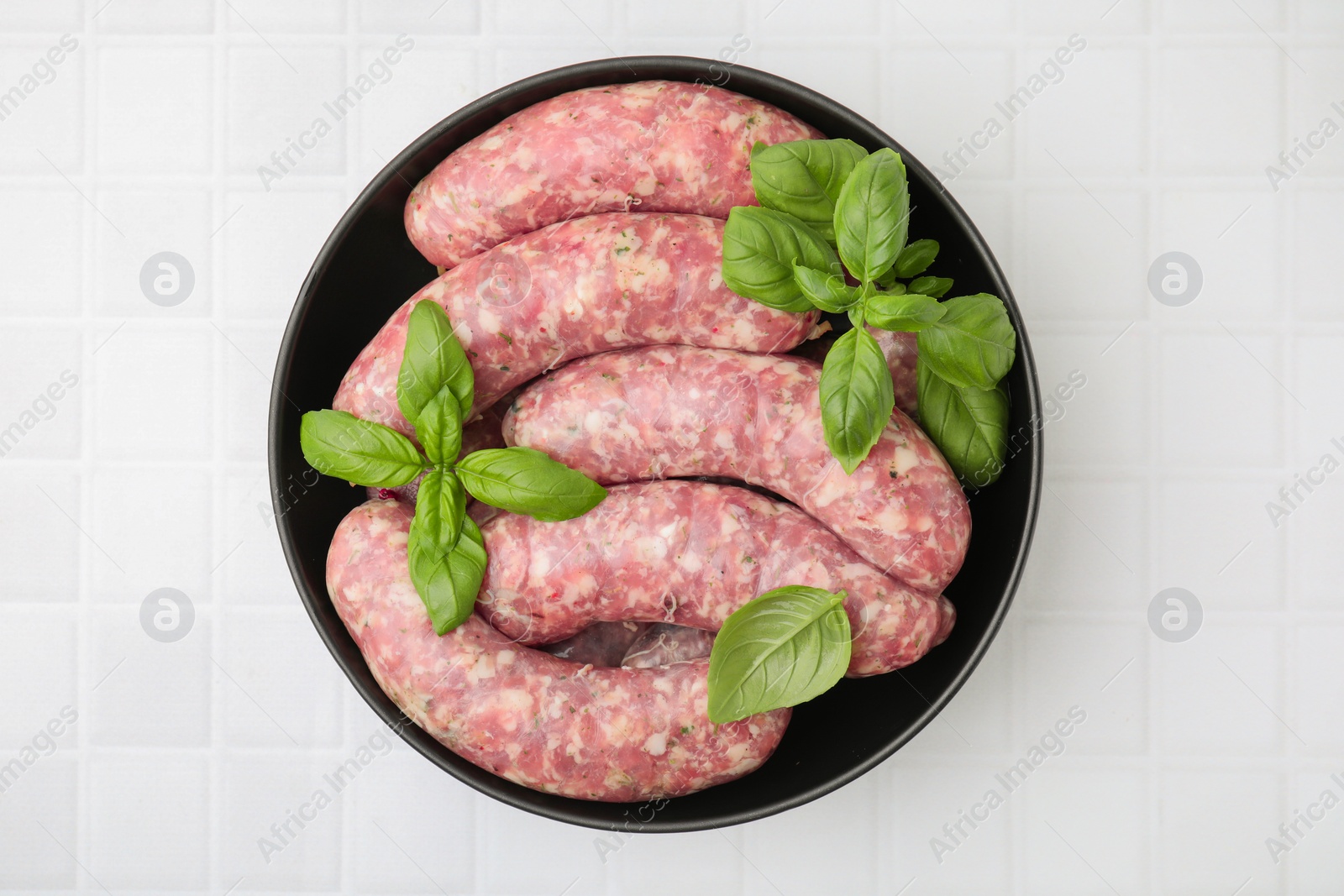 Photo of Raw homemade sausages and basil leaves on white tiled table, top view