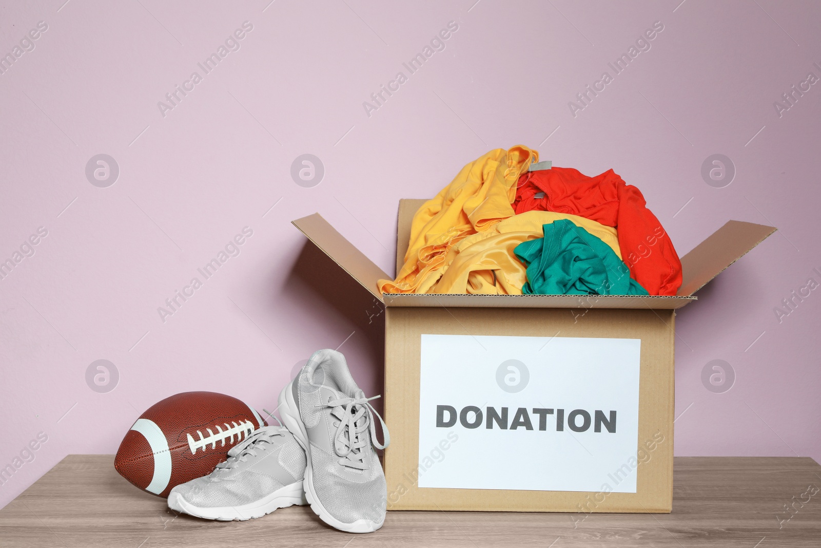 Photo of Donation box with clothes, shoes and rugby ball on table against color background