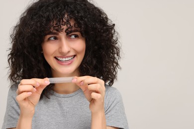Photo of Young woman holding teeth whitening strip on light grey background, space for text